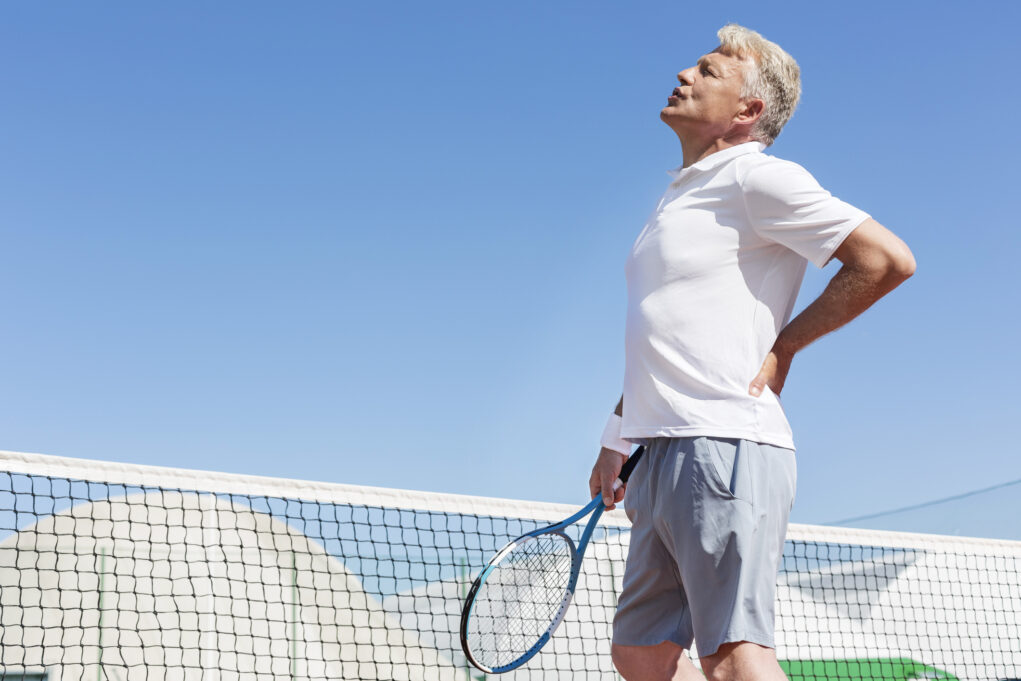 Mature man grimacing with backache while holding tennis racket against clear blue sky on sunny day
